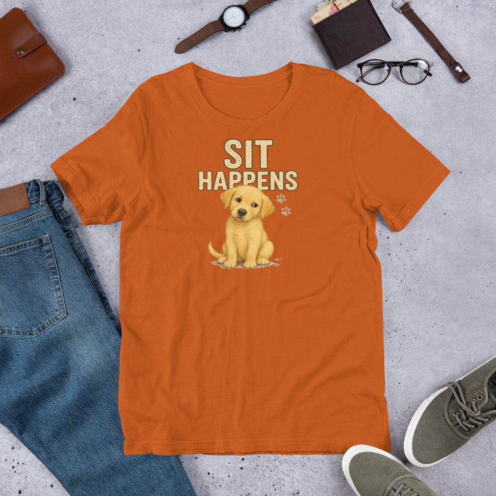 "Sit Happens " – Fun Labrador T-Shirt Orange t-shirt with a puppy graphic and 'Sit Happens' text on a concrete surface with casual items.