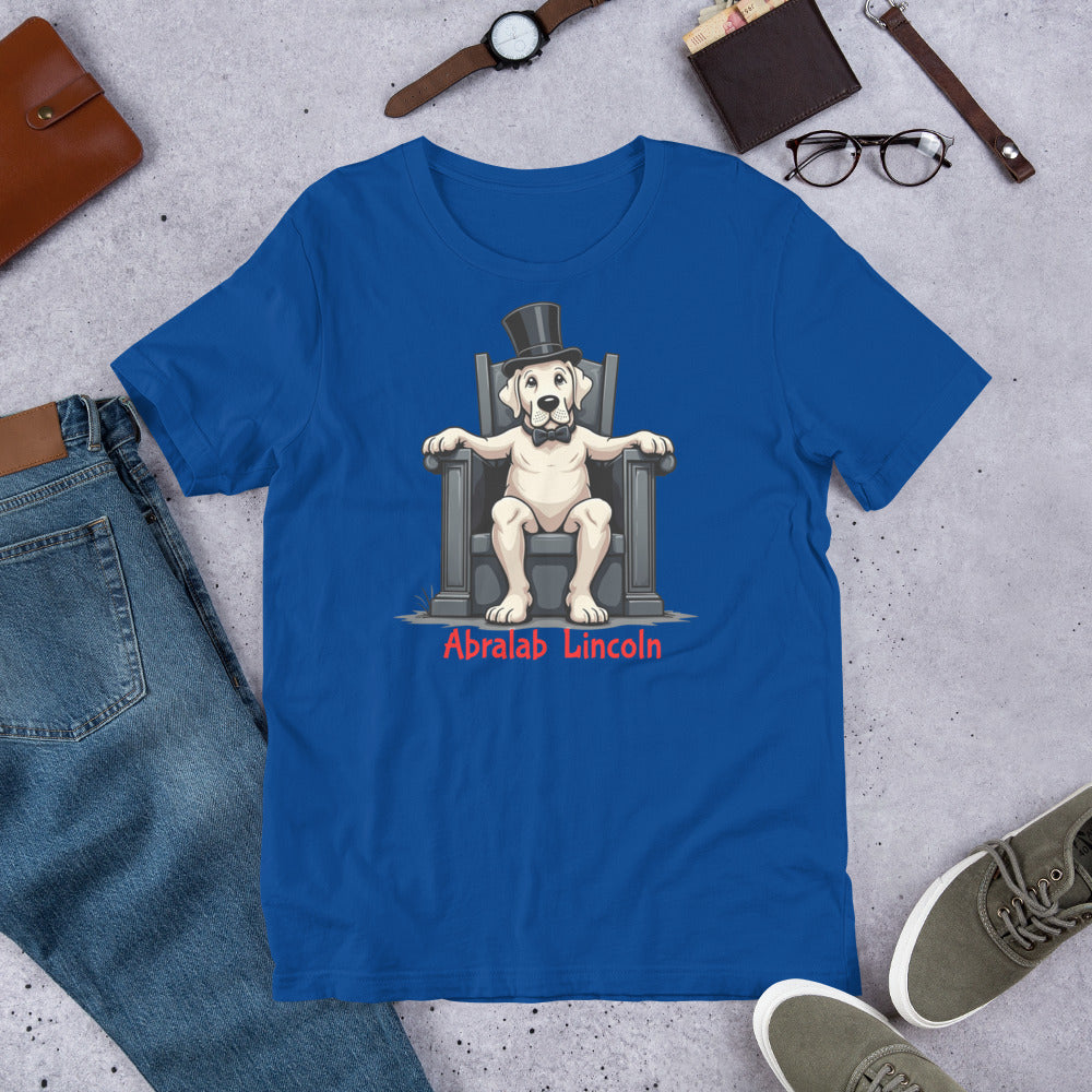 Blue t-shirt with a dog wearing a top hat and monocle design, surrounded by jeans, shoes, and accessories on a concrete surface. T-shirt with bold black text that says “Abralab Lincoln,” styled flat with jeans, shoes, and a hat. A playful Labrador lover shirt and perfect gift for Labrador owners who appreciate humor.