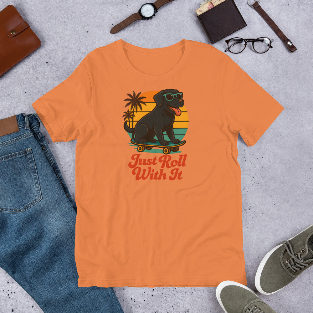 "Just Roll With It" - Black Labrador – Fun Labrador T-Shirt Orange t-shirt with a graphic of a dog on a skateboard and text 'Just Roll With It' displayed on a concrete surface with casual items.