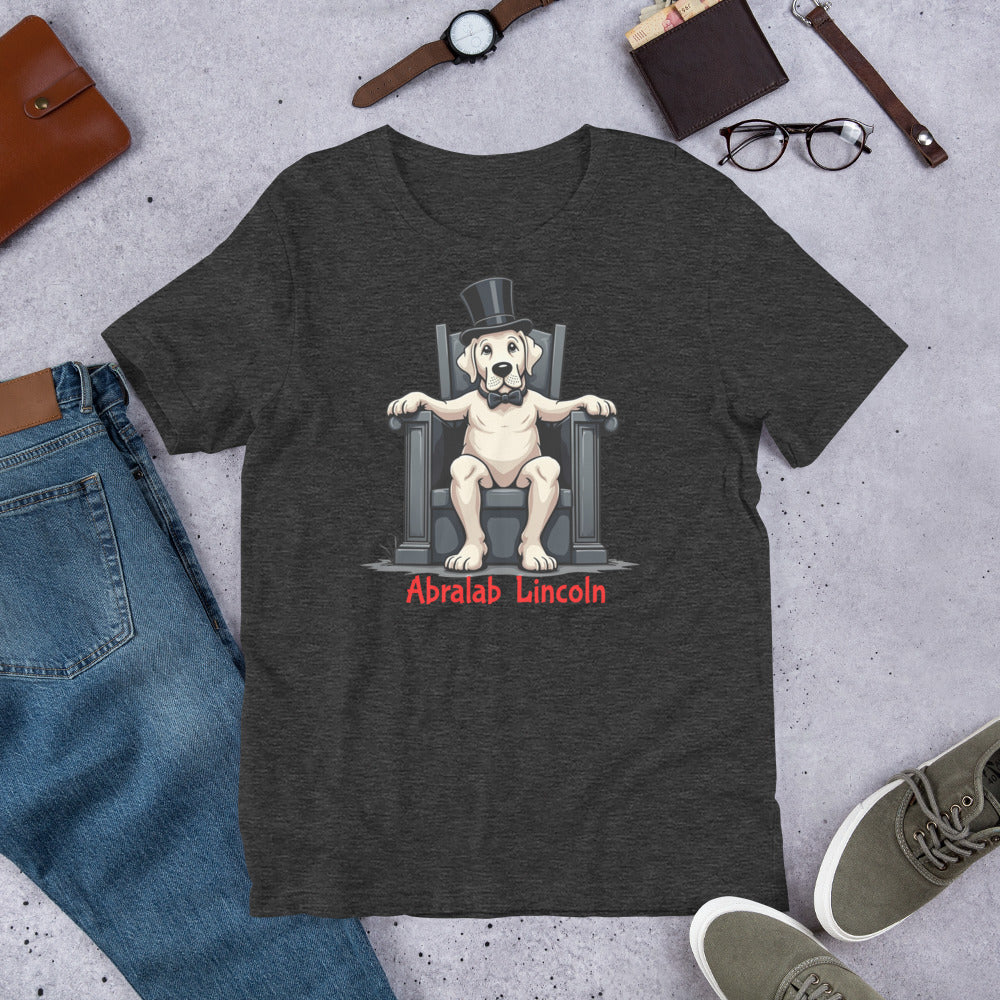Dark gray t-shirt with a graphic of a dog in a top hat and bow tie sitting on a throne, labeled 'Abralab Lincoln', displayed with jeans, shoes, and accessories. T-shirt with bold black text that says “Abralab Lincoln,” styled flat with jeans, shoes, and a hat. A playful Labrador lover shirt and perfect gift for Labrador owners who appreciate humor.