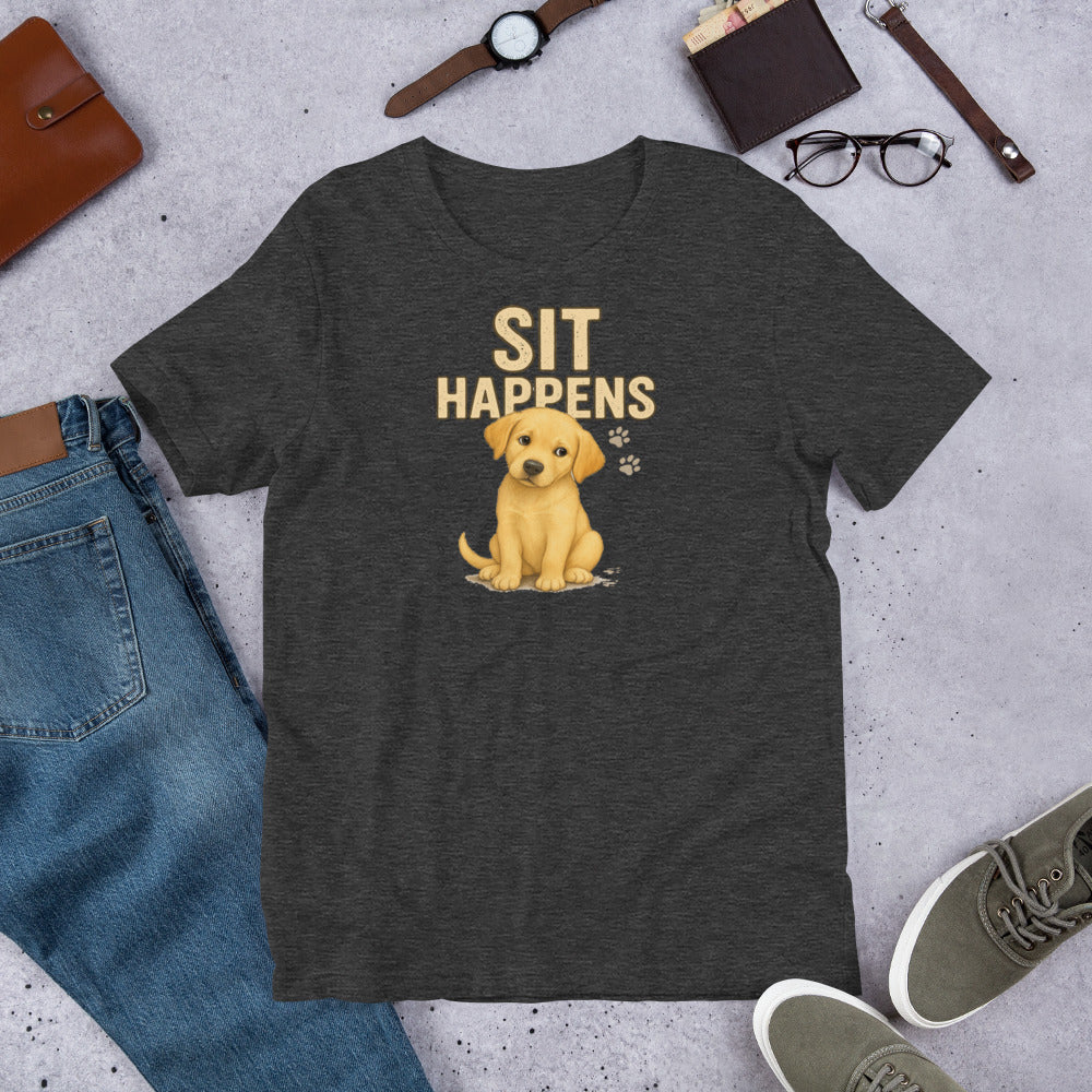 "Sit Happens " – Fun Labrador T-Shirt Dark gray t-shirt with a yellow puppy graphic and 'Sit Happens' text, surrounded by jeans, shoes, and accessories on a concrete surface.