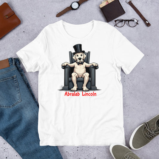 White t-shirt with a dog wearing a top hat and bow tie design, surrounded by jeans, shoes, and accessories on a gray surface. T-shirt with bold black text that says “Abralab Lincoln,” styled flat with jeans, shoes, and a hat. A playful Labrador lover shirt and perfect gift for Labrador owners who appreciate humor.