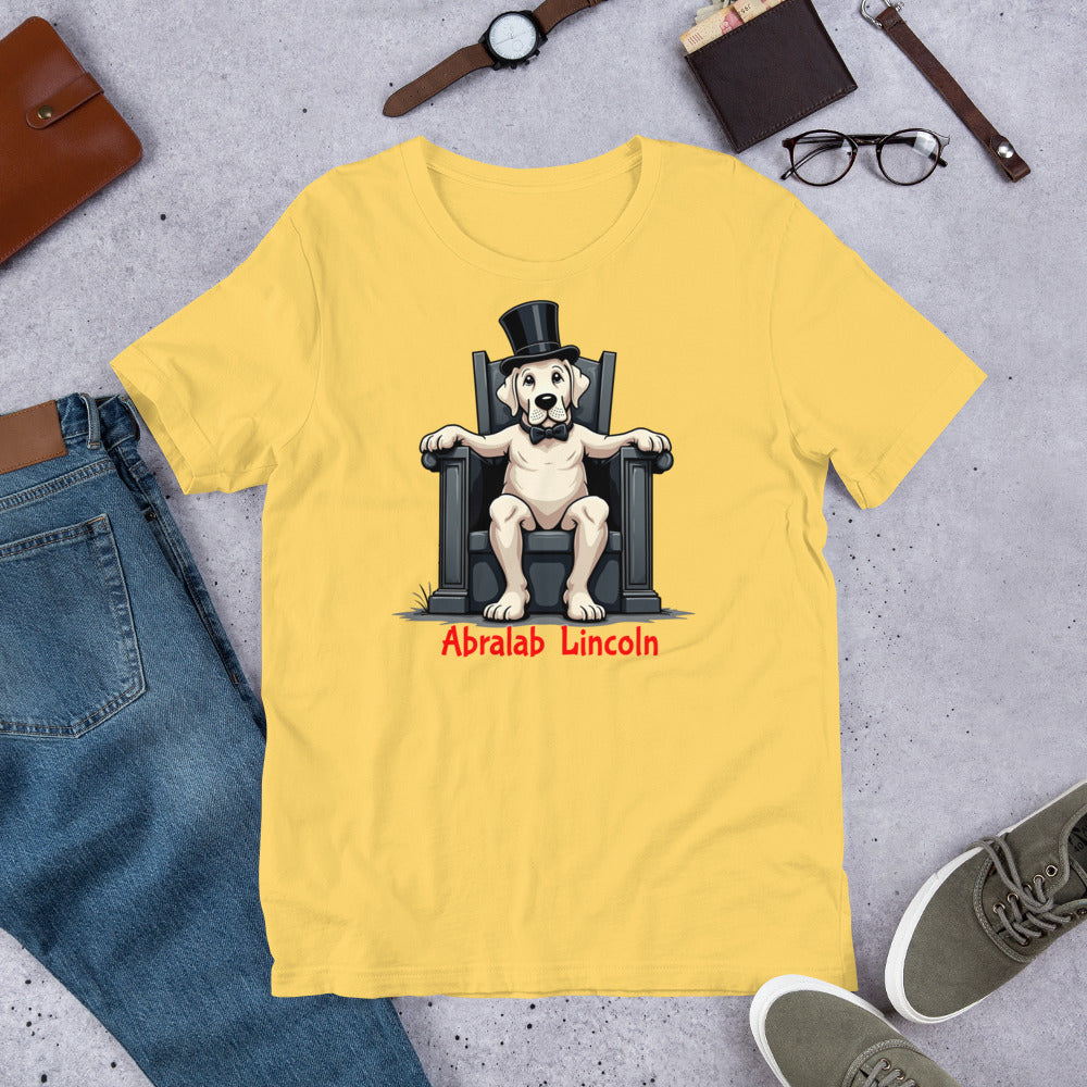 Yellow t-shirt with a dog wearing a top hat and monocle design, surrounded by jeans, shoes, and accessories on a concrete surface. T-shirt with bold black text that says “Abralab Lincoln,” styled flat with jeans, shoes, and a hat. A playful Labrador lover shirt and perfect gift for Labrador owners who appreciate humor.